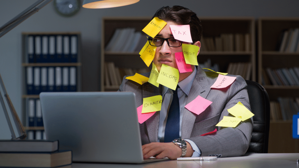 Picture of an SMB leader sitting at his desk in front of his laptop with a lot of post it notes stuck all over him indicating the competing priorities he has. The post-its have words like "dinner", "income", "tax", "expenses", and "report" labelled on them.