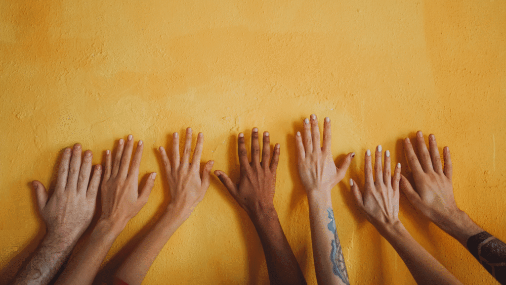 Photo of a series of hands which appear to be diverse in many ways, raised together against a yellow wall.