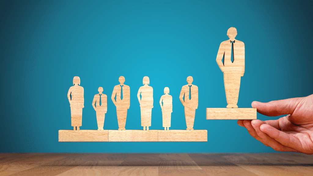 Picture of a hand placing a wooden cutout of a business person beside six other wooden cutouts. All the cutouts have a wood base that join together as a team as they hover over the desk.