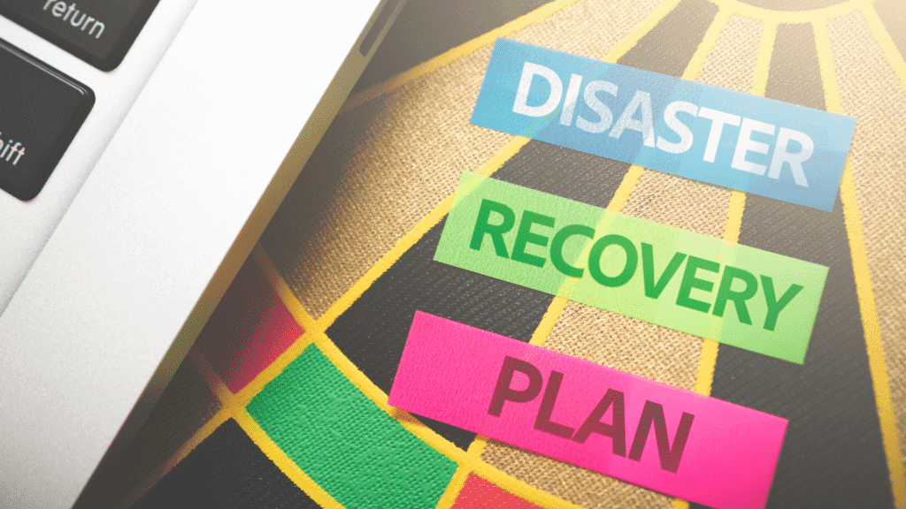 Abstract picture of the side of a laptop sitting on top of what appears to be a dart board. On the dart board are words on tape. The inner tape line says "Disaster". Then "Recovery". Then "Plan".