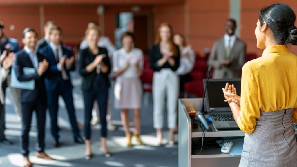 A woman presents her team's success in delivering value to the team in a stand-up meeting. The team is applauding their work.